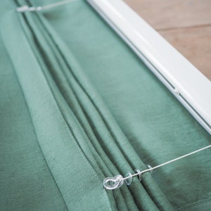 K&ouml;nnte beinhalten: Nahaufnahme eines gr&uuml;nen Stoff-Rollos mit einer wei&szlig;en Schiene und transparenten Kunststoffringen. Das Rollo ist hochgezogen und die Ringe sind sichtbar.