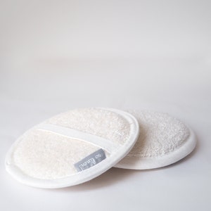 May include: Two white round bath poufs with a white trim and a gray label that says "vanity co."