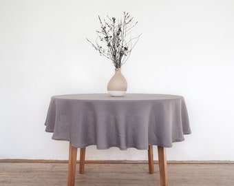 Puede incluir: Un mantel redondo de lino gris drapeado sobre una mesa de madera con un jarrón de ramas secas en el centro.