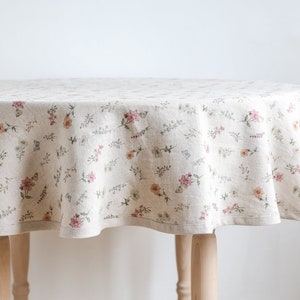 Peut inclure: Une nappe ronde de couleur crème avec un motif floral délicat dans des tons de rose, vert et jaune. La nappe est en lin et a un léger brillant.
