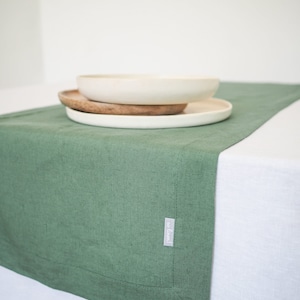 Può includere: Un runner da tavolo in lino verde con un'etichetta bianca che dice "eco linen". Il runner è su una tovaglia bianca e ci sono tre piatti impilati uno sopra l'altro.