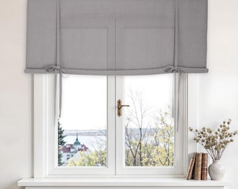 Puede incluir: Una cortina romana gris está atada sobre una ventana blanca. La ventana tiene un tirador dorado y una vista de un lago y edificios. Un jarrón con flores secas y libros están en el alféizar.