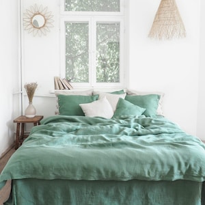 May include: A bed with a green linen duvet cover and matching pillows. The room has a neutral color palette with a wooden side table, a decorative mirror, and a woven pendant light. The bed is the focal point of the room.