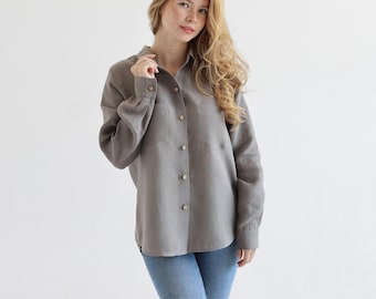 May include: A gray linen shirt with long sleeves and a button-down front. The shirt has a chest pocket and is worn with blue jeans.