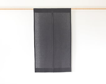 Puede incluir: Un panel de cortina de lino gris oscuro colgado de una barra de madera sobre un fondo blanco. El panel es rectangular con una apariencia texturizada y un borde más oscuro. La tela está doblada por la mitad.