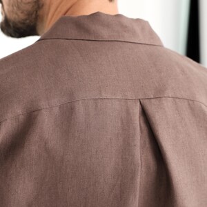 May include: Close-up of the back of a brown button-down shirt. The shirt is made of a textured fabric and features a collar and a pleat detail at the back yoke. The color is a muted, earthy tone.