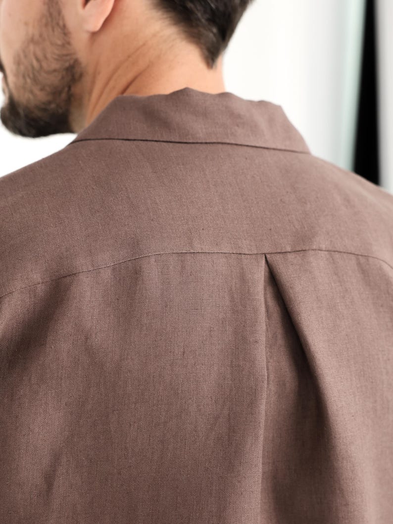 May include: Close-up of the back of a brown button-down shirt. The shirt is made of a textured fabric and features a collar and a pleat detail at the back yoke. The color is a muted, earthy tone.