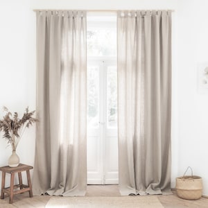 May include: A pair of beige linen curtains hang from a wooden rod in a room with white walls. The curtains are gathered at the top and hang loosely, creating a relaxed and casual look.