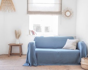 Może przedstawiać: Jasnoniebieska sofa z narzutą i poduszką. Sofa znajduje się w salonie z drewniana podłogą, małym drewnianym stolikiem i oknem z białą roletą.