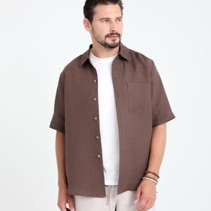 May include: A short-sleeved, button-down shirt in a rich brown color. The shirt has a single chest pocket and is worn over a white t-shirt. The model is wearing beige pants and a bracelet. The background is plain white.