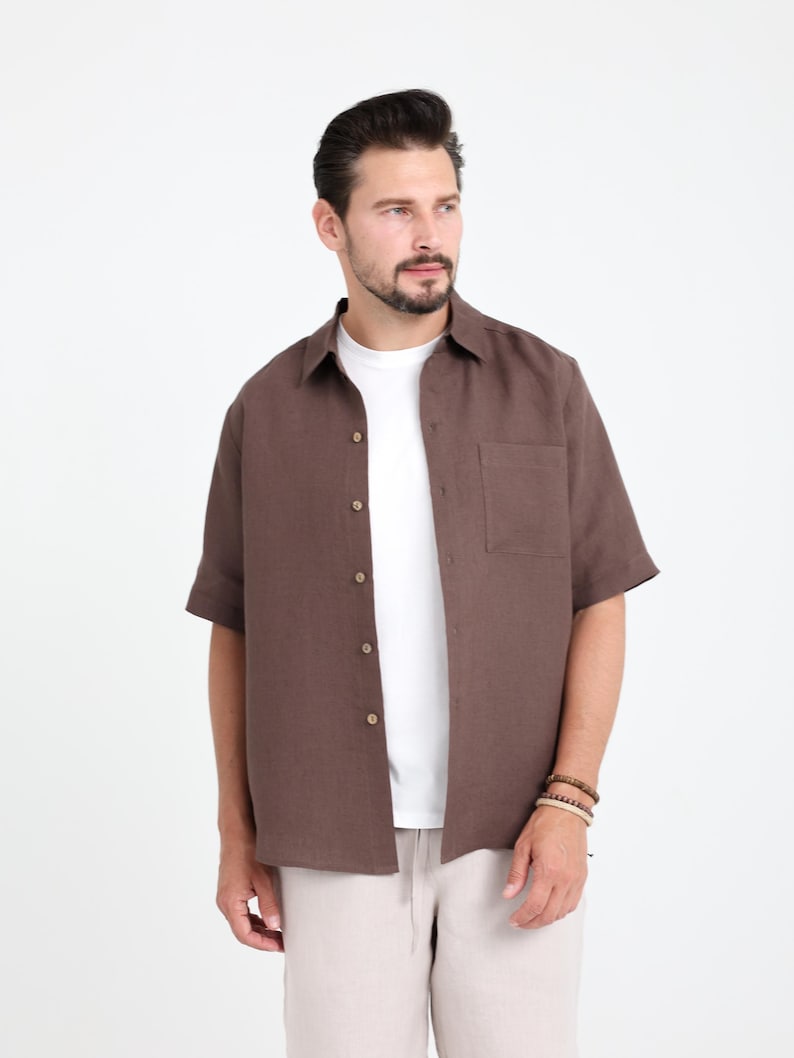 May include: A short-sleeved, button-down shirt in a rich brown color. The shirt has a single chest pocket and is worn over a white t-shirt. The model is wearing beige pants and a bracelet. The background is plain white.