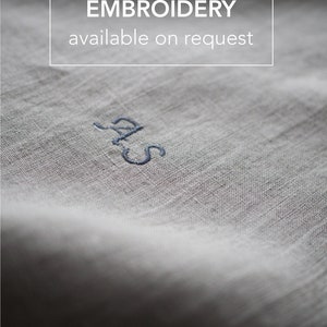 Op de afbeelding: Close-up van een witte linnen stof met de tekst "EMBROIDERY available on request" en de initialen "S.A.S." geborduurd in blauwe draad.