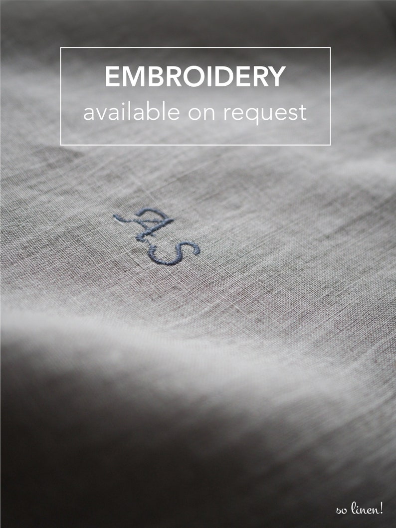 Op de afbeelding: Close-up van een witte linnen stof met de tekst "EMBROIDERY available on request" en de initialen "S.A.S." geborduurd in blauwe draad.