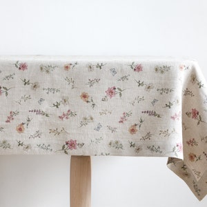 Puede incluir: Un mantel de lino beige con un delicado estampado floral con flores rosas, amarillas y verdes y mariposas.