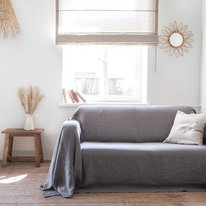 Könnte beinhalten: Ein graues Leinen-Sofa mit einer Überwurfdecke und einem Kissen. Das Sofa steht in einem Wohnzimmer mit einem Fenster, einem Beistelltisch und einer Hängeleuchte aus Weide.