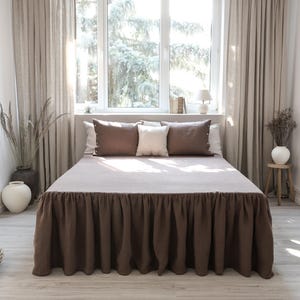 May include: A bed with a light brown bedspread and a dark brown bed skirt. The bed has three pillows, two brown and one white. The room has beige curtains, a straw hat, and a woven basket. A vase of dried flowers sits on a small wooden stool.