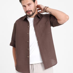 May include: A brown short-sleeve button-down shirt with a chest pocket. The shirt is worn over a white t-shirt and paired with light-colored pants. The model is wearing bracelets on their wrist.