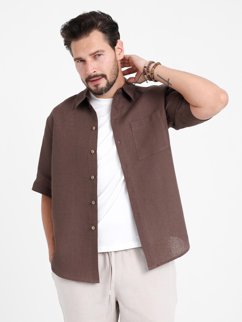 May include: A brown short-sleeve button-down shirt with a chest pocket. The shirt is worn over a white t-shirt and paired with light-colored pants. The model is wearing bracelets on their wrist.