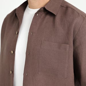May include: A brown button-down shirt with a chest pocket. The shirt is made of a textured fabric and has a classic collar. A white t-shirt is visible underneath the brown shirt.