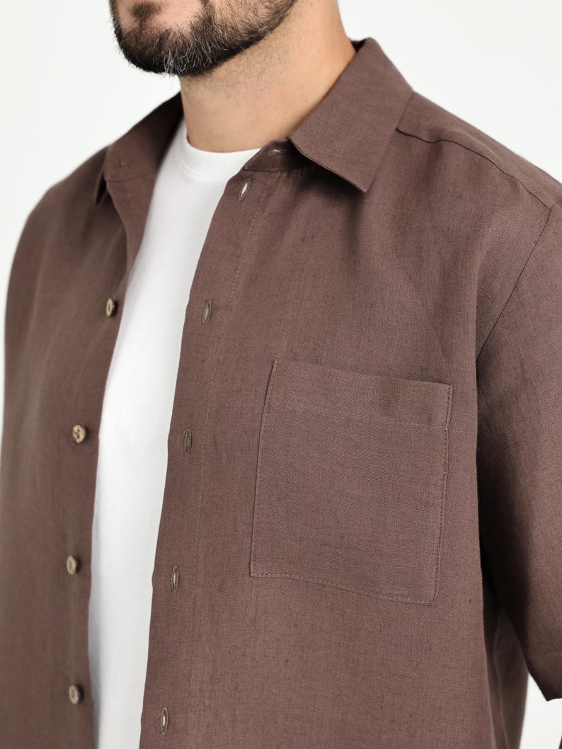 May include: A brown button-down shirt with a chest pocket. The shirt is made of a textured fabric and has a classic collar. A white t-shirt is visible underneath the brown shirt.