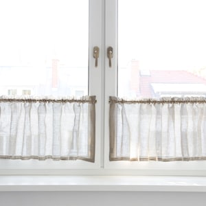 Op de afbeelding: Twee kleine, lichtgekleurde linnen café gordijnen hangen aan een wit raam. De gordijnen zijn aan de bovenkant geplooid en hebben een eenvoudig, neutraal ontwerp. Het raam is helder, met uitzicht op daken en lucht.