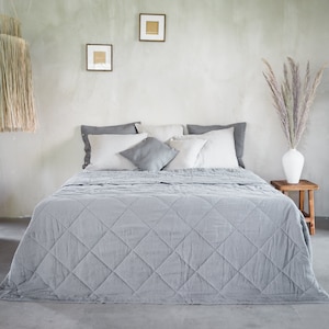 Könnte beinhalten: Eine graue Leinen-Steppdecke mit Rauten-Steppung auf einem Bett mit weißen Leinenkissen. Das Bett steht in einem Raum mit einer Betonwand und einer Vase mit Trockenblumen.