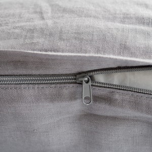 Puede incluir: Una almohada de lino gris con cierre de cremallera. La cremallera es visible y está hecha de metal.