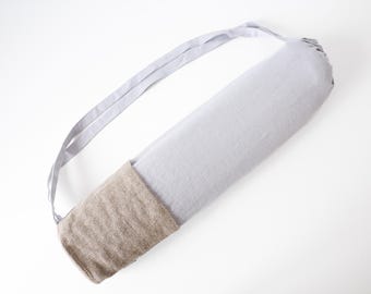 May include: A light gray yoga mat bag with a matching shoulder strap. The bottom of the bag is a tan, textured fabric. The bag is designed to carry a rolled-up yoga mat.