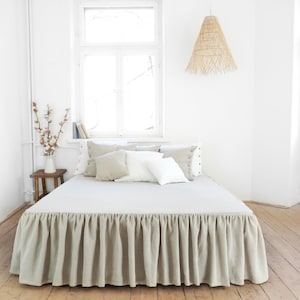 Può includere: Una gonna da letto in lino beige con un bordo arricciato, su un letto bianco con cuscini bianchi e beige in una camera da letto con una parete bianca e una finestra.