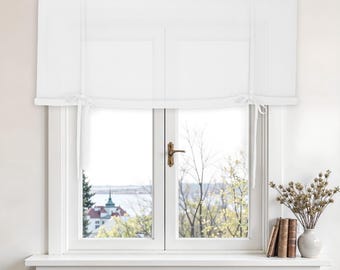 Puede incluir: Ventana luminosa con marco blanco y cortina romana blanca. Vista de un paisaje con agua y edificios. Un jarrón con flores secas y libros en el alféizar crean una escena luminosa y aireada.