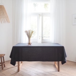 Puede incluir: Un mantel de lino negro drapeado sobre una mesa de madera con un jarrón de flores secas en el centro. La mesa está puesta en una habitación con paredes blancas y grandes ventanas con cortinas blancas.