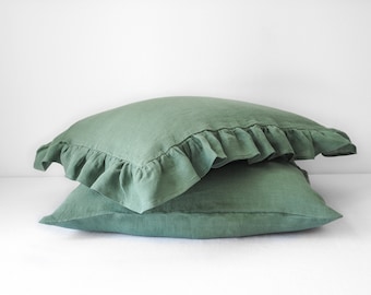Puede incluir: Dos fundas de almohada de lino verde salvia. La funda de almohada superior tiene un borde con volantes. Las almohadas están apiladas. El fondo es blanco.