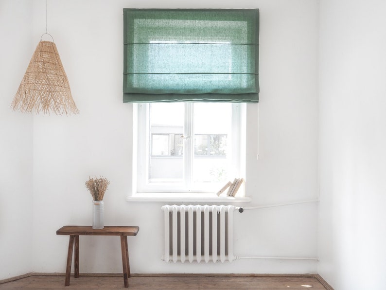 K&ouml;nnte beinhalten: Ein wei&szlig;er Raum mit einem gr&uuml;nen Leinen-Rollos an einem Fenster. Das Fenster hat einen wei&szlig;en Rahmen und einen wei&szlig;en Heizk&ouml;rper darunter. Es gibt einen Holzhocker und eine gewebte Lampenfassung, die von der Decke h&auml;ngt.