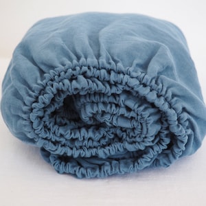 Op de afbeelding: Een opgerold, blauw hoeslaken. Het laken is gemaakt van een zachte, getextureerde stof en heeft elastische randen. De kleur is een gedempt, stoffig blauw, en het laken lijkt nieuw en ongebruikt. Het laken is ontworpen voor een bed.