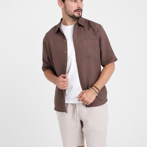 May include: A brown short-sleeved shirt with a chest pocket, worn over a white t-shirt, paired with beige shorts. The outfit is casual and suitable for warm weather. The model is wearing bracelets.