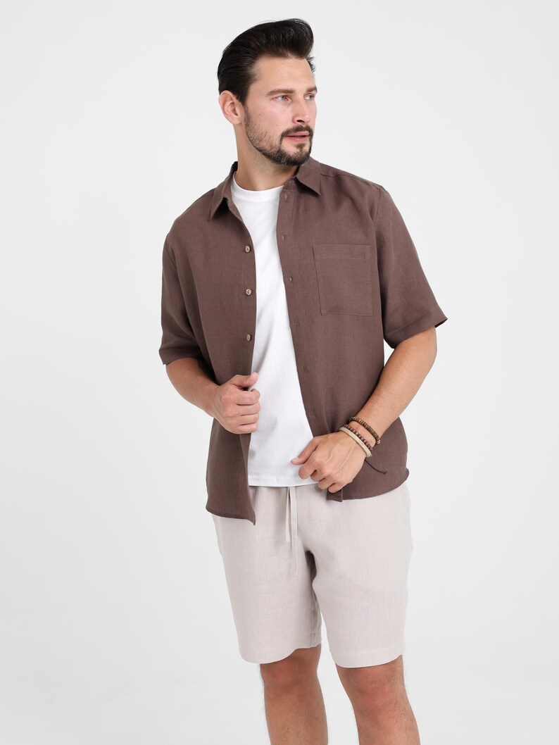 May include: A brown short-sleeved shirt with a chest pocket, worn over a white t-shirt, paired with beige shorts. The outfit is casual and suitable for warm weather. The model is wearing bracelets.
