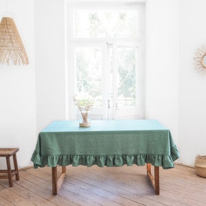May include: A sage green linen tablecloth with a ruffled edge, draped over a wooden table. The tablecloth is set in a room with a white wall, a wooden floor, and a window with natural light.