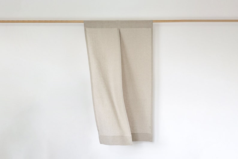 May include: A pair of beige linen curtains hanging from a wooden rod. The curtains have a subtle texture and a slightly darker border.