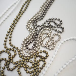 K&ouml;nnte beinhalten: Drei verschiedene Arten von Perlenketten, eine in Gold, eine in Silber und eine in Schwarz und Silber. Die Ketten liegen alle auf einer wei&szlig;en Oberfl&auml;che.