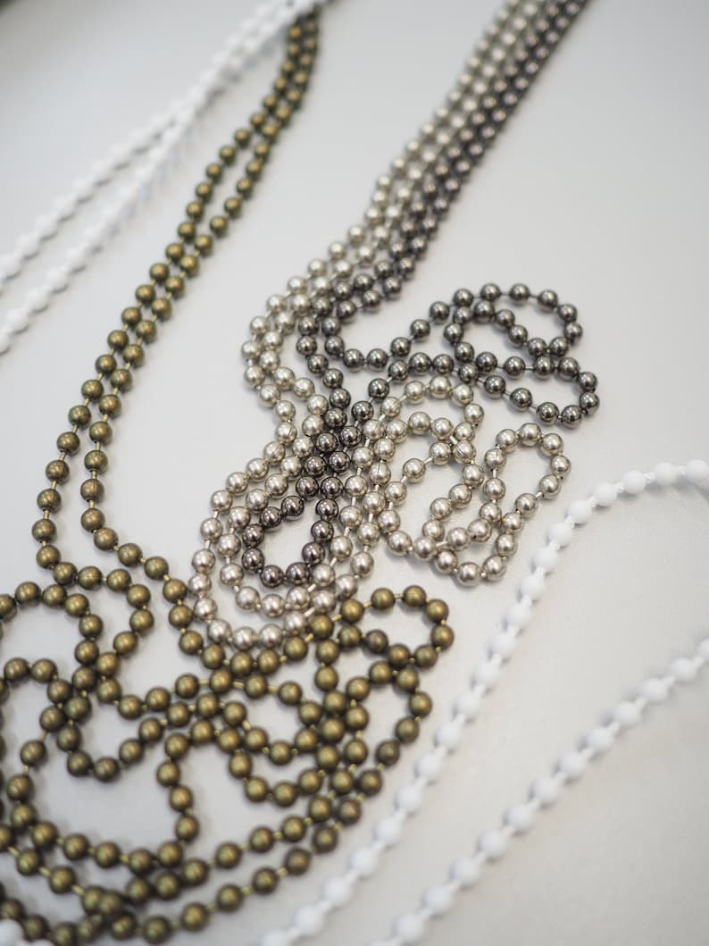 K&ouml;nnte beinhalten: Drei verschiedene Arten von Perlenketten, eine in Gold, eine in Silber und eine in Schwarz und Silber. Die Ketten liegen alle auf einer wei&szlig;en Oberfl&auml;che.