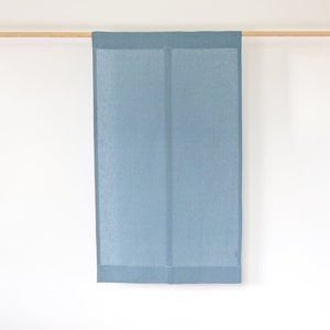 Pode incluir: Uma cortina ou painel retangular de linho azul claro pende de uma barra de madeira sobre um fundo branco. O tecido tem uma textura subtil e uma borda com bainha. A cortina é provavelmente usada para revestimentos de janelas ou divisórias de ambientes.