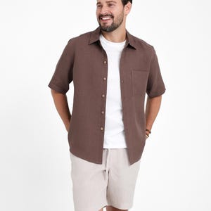May include: A short-sleeved brown button-down shirt with a chest pocket, worn over a white t-shirt. Paired with beige shorts. The shirt has a classic collar and wooden buttons. The outfit is casual and relaxed.
