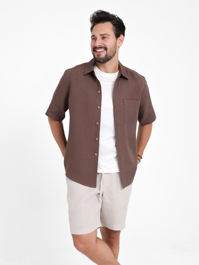 May include: A short-sleeved brown button-down shirt with a chest pocket, worn over a white t-shirt. Paired with beige shorts. The shirt has a classic collar and wooden buttons. The outfit is casual and relaxed.