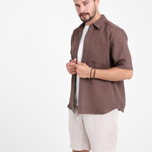 May include: A brown short-sleeved shirt with a chest pocket, worn over a white t-shirt, paired with beige shorts. The outfit is completed with a wooden beaded bracelet. The clothing is casual and suitable for warm weather.