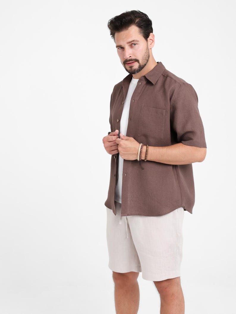 May include: A brown short-sleeved shirt with a chest pocket, worn over a white t-shirt, paired with beige shorts. The outfit is completed with a wooden beaded bracelet. The clothing is casual and suitable for warm weather.
