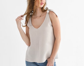 May include: A light beige linen tank top with thin straps that tie at the shoulders. The top has a v-neck and a relaxed fit. The model is wearing blue jeans.