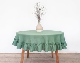 May include: A round, sage green linen tablecloth with a ruffled edge, draped over a wooden-legged table. A beige vase with dried stems sits in the center. The table is set against a white background.