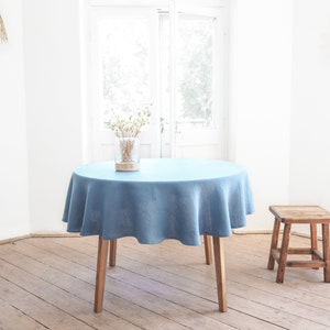 Op de afbeelding: Een ronde houten tafel met een lichtblauw linnen tafelkleed. De tafel staat in een kamer met witte muren en houten vloer. Een kleine houten kruk staat op de voorgrond.