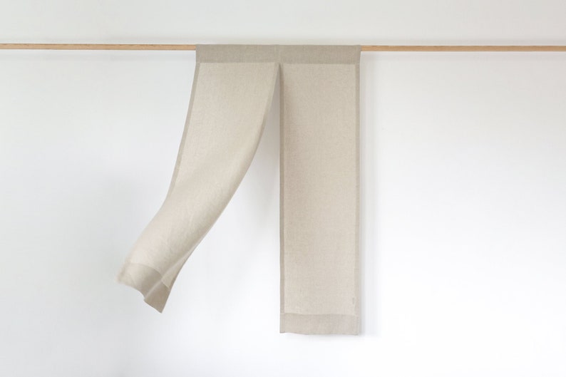 May include: A beige linen scarf with a fringed edge hangs from a wooden rod against a white wall.