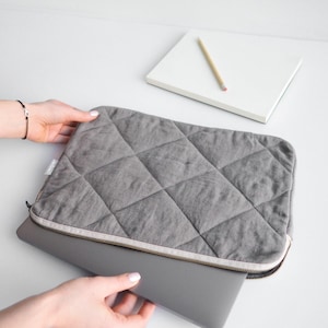 Könnte beinhalten: Eine graue, gesteppte Laptoptasche mit Reißverschluss. Die Tasche ist offen und ein Laptop befindet sich teilweise im Inneren.